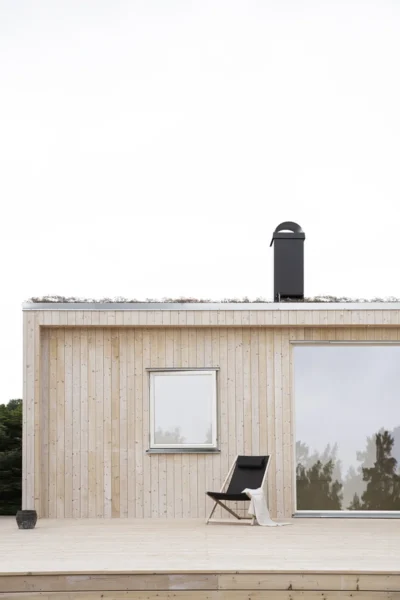 Detaljbild på ett trähus med altan framför. På altanen står en fåtölj. Huset har två fönster och en svart skorsten. Taket är ett sedumtak och man ser växterna sticka upp ovanför en kantlist i zink.