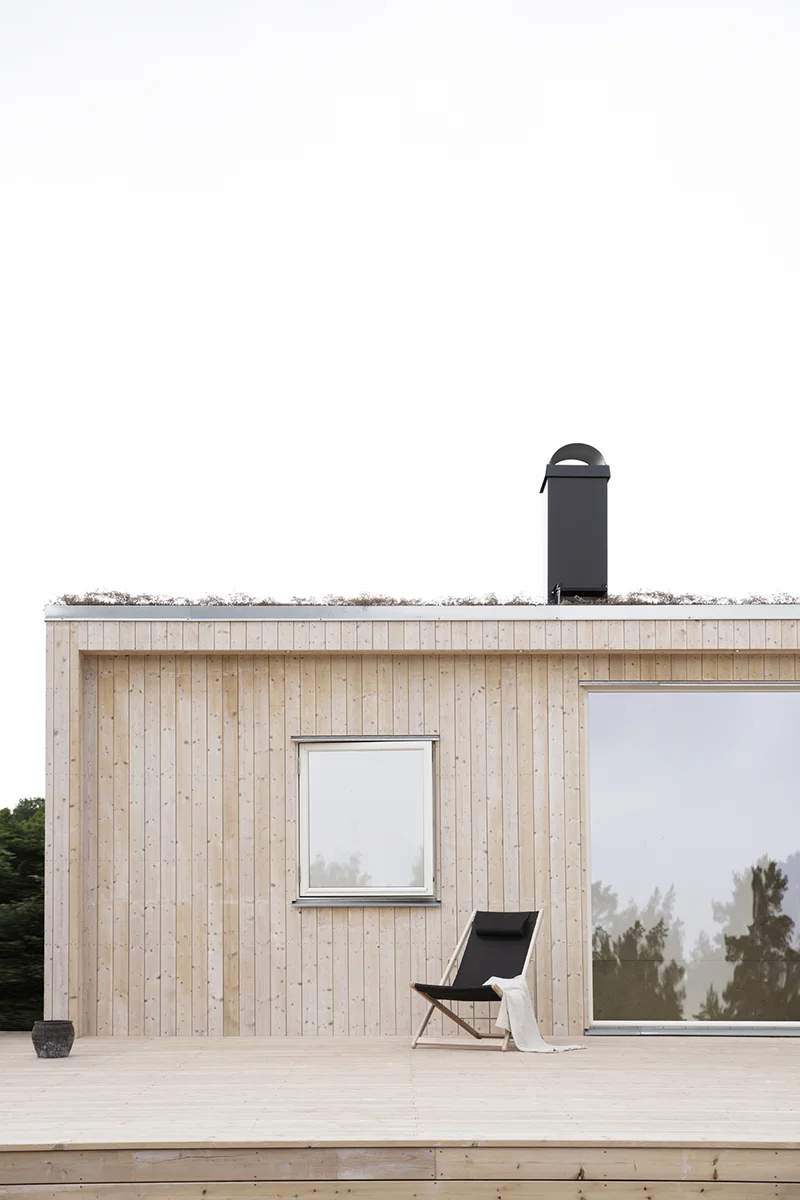 Detaljbild på ett trähus med altan framför. På altanen står en fåtölj. Huset har två fönster och en svart skorsten. Taket är ett sedumtak och man ser växterna sticka upp ovanför en kantlist i zink.