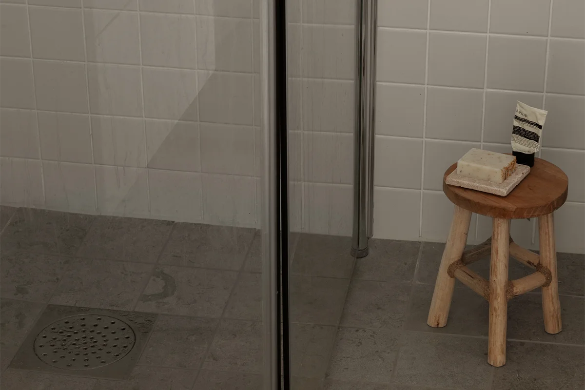 Duschhörna med duschväggar, ljusbeige kakel och mörkare klinkergolv. Bredvid duschen står en träpall med tvål på ett tvålfat och en tub handkräm.