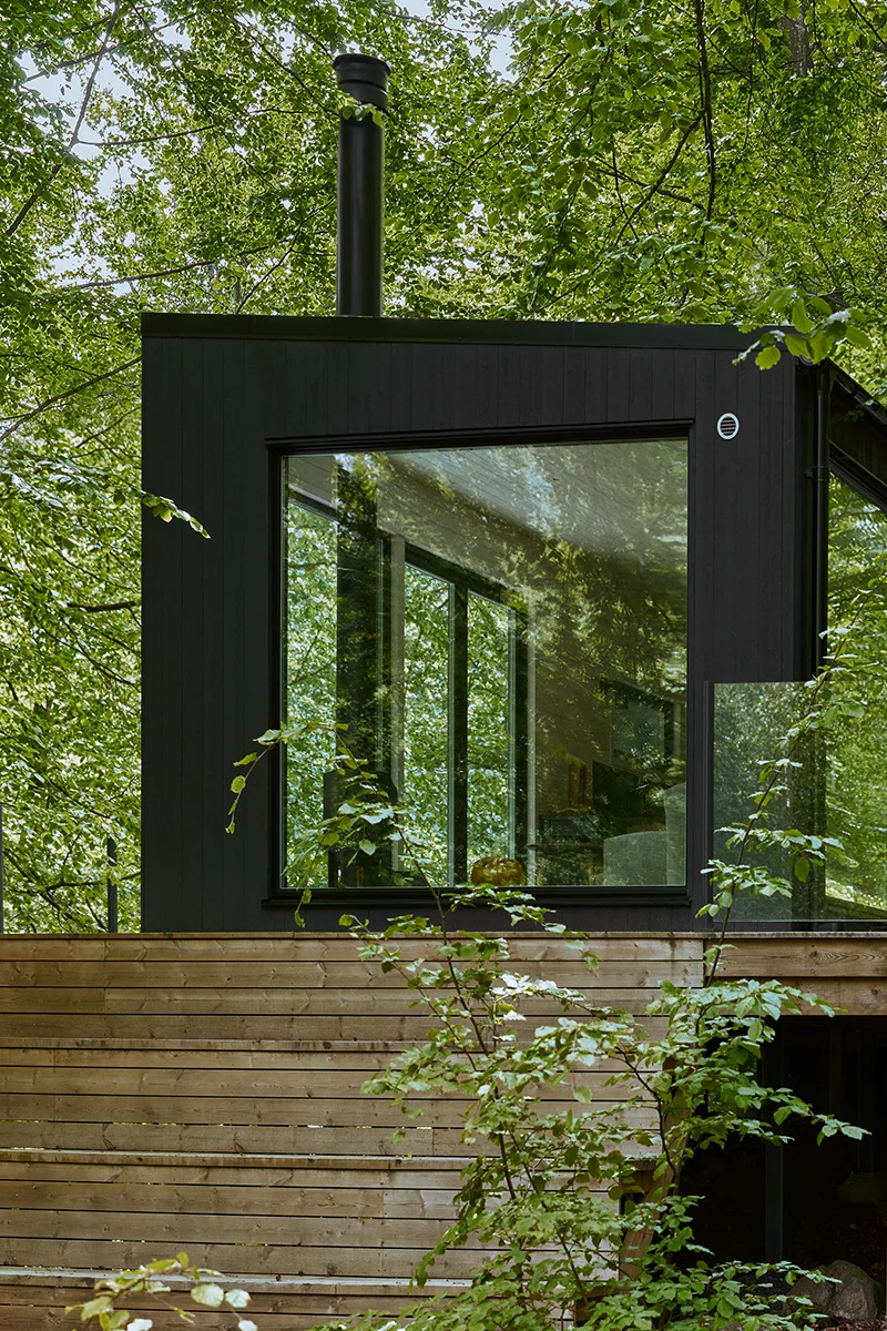 Svart trähus med Svanenmärkta träfönster. Huset står på en träaltan med trappa upp till gaveln på huset, där ett stort fönster sitter över nästan hela väggen. Huset är omringat av träd med gröna löv.
