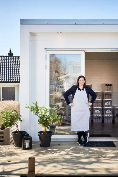 Kvinna står på en altan med krukväxter framför ett vitt attefallshus med fönsterdörr