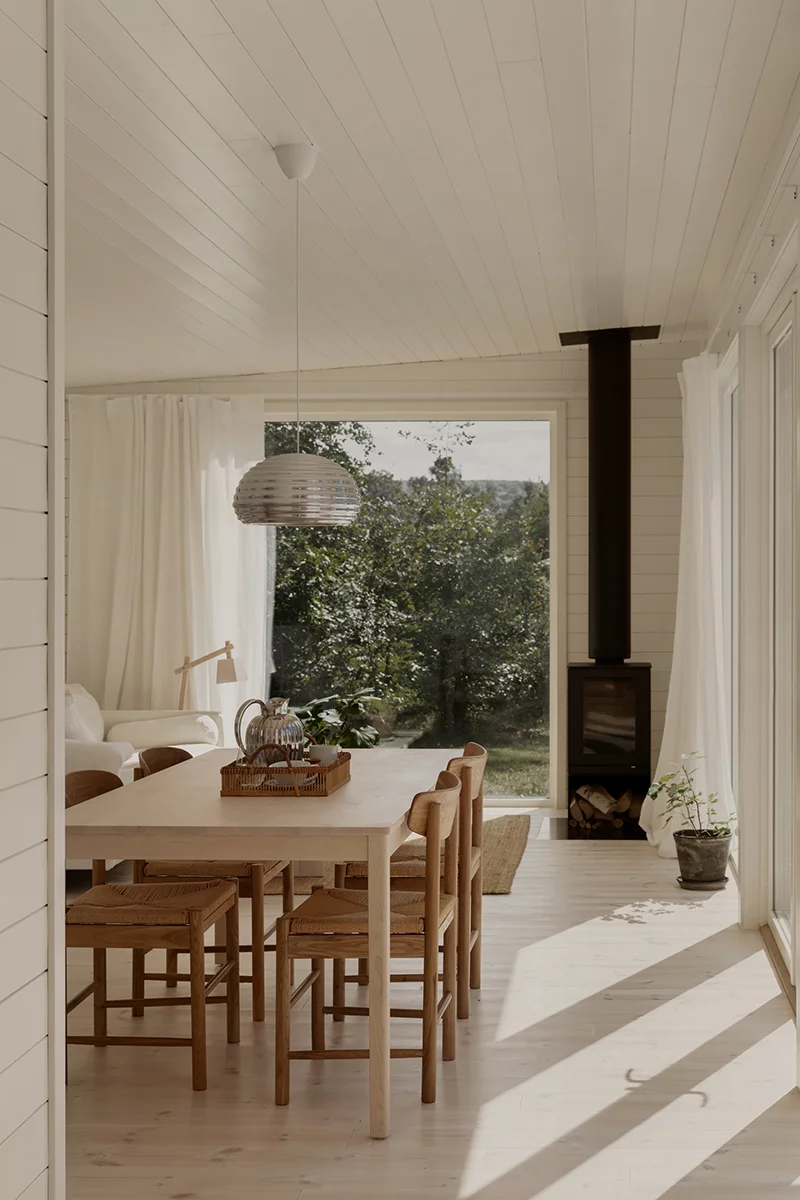 Vitmålad träpanel i visningshuset i Båstad, inrett med matplats och kamin framför ett gavelfönster