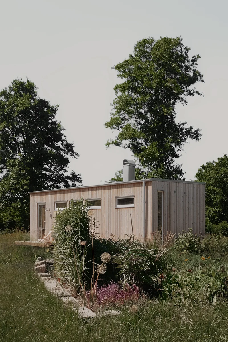 Exteriör i ljus träpanel på visningshuset på Resö, omgivet av träd, blommor och växter