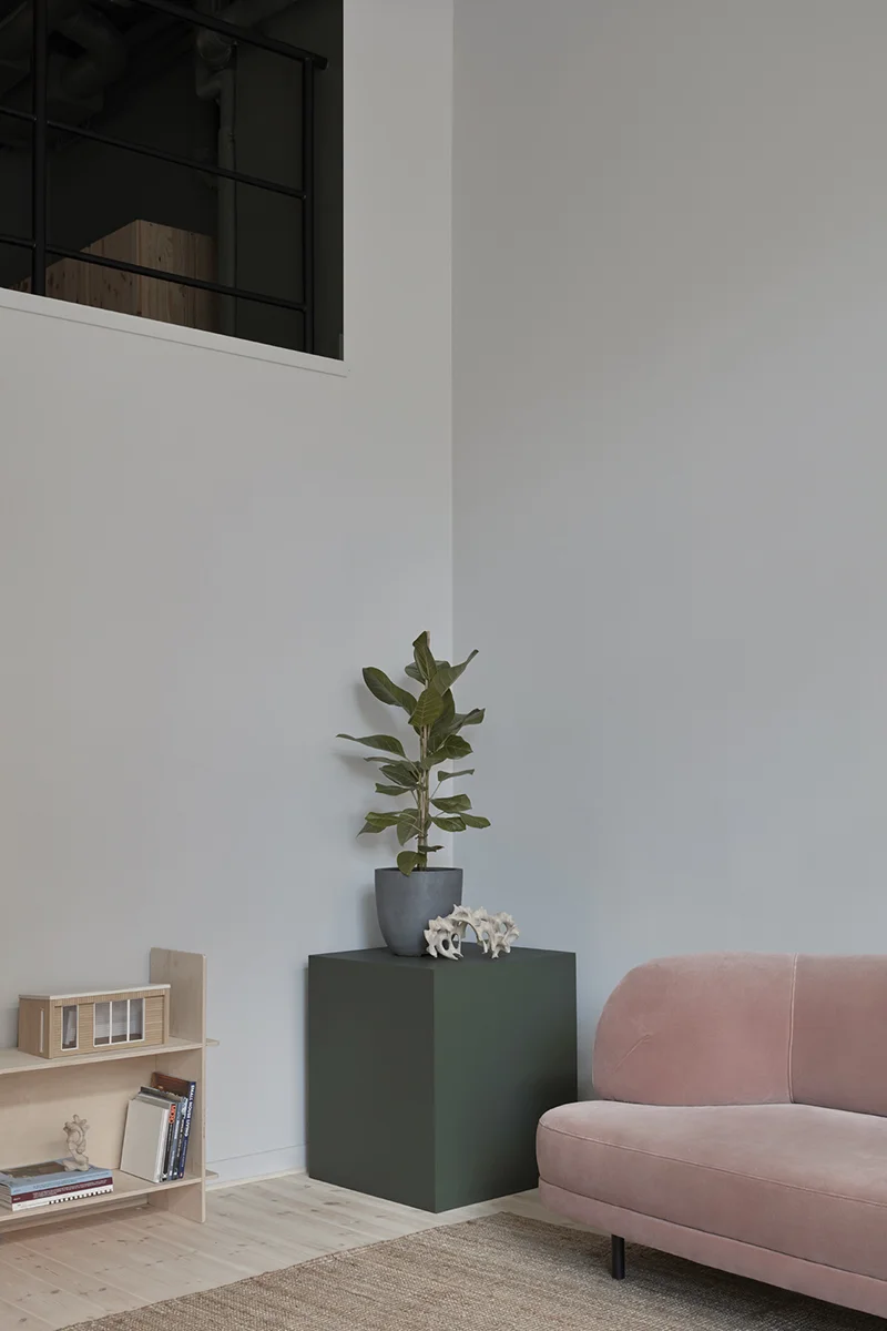 Hörn med lite bokhylla, piedestal med växt, rosa soffa på beige matta