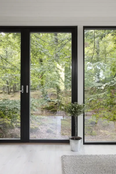 Interiør med hvitmalte vegger, hvitt vokset gulv og svarte vinduer med utsikt over en grønn skog