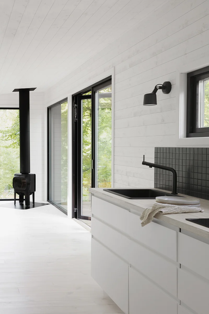 Kjøkken i Enkelrum Nest åpner seg mot stuen med komfyr, interiøret har hvitmalt trepanel.